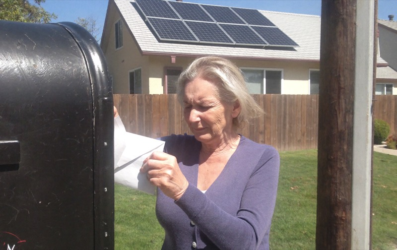 A homeowner pulls a utility bill from the mailbox in front of a house with solar panels